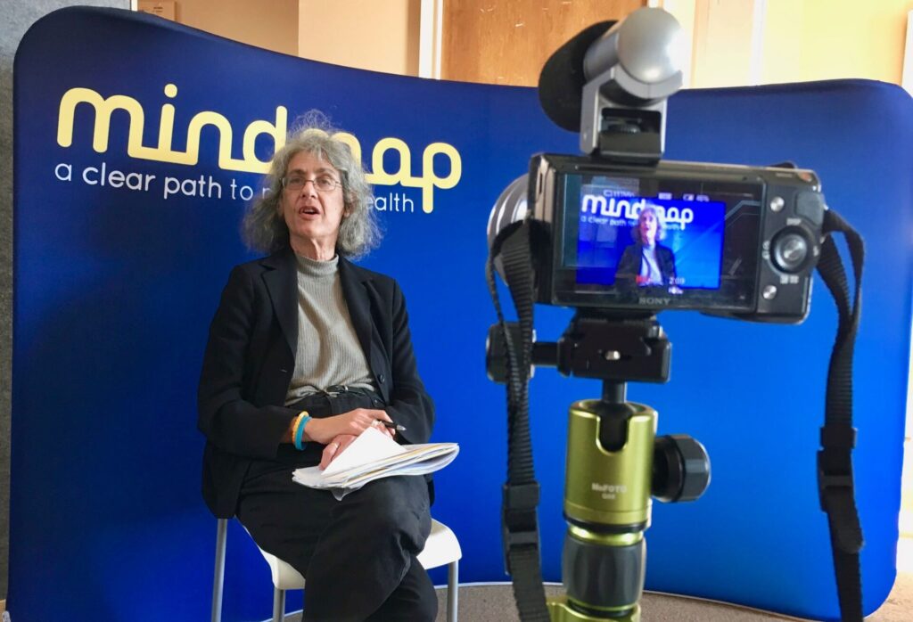 Ellen Saks in front of a mindmap backdrop filming an interview